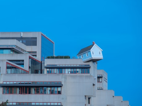 A unique architectural scene with a small white house perched precariously on the edge of a modern concrete building. The building has multiple levels with a minimalist design and red trim around the windows, set against a clear blue sky.