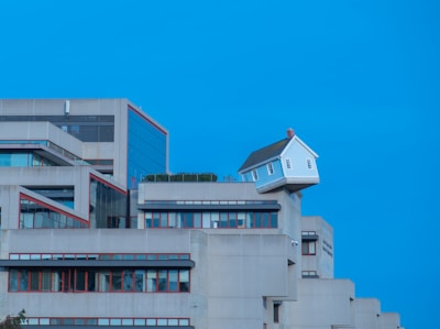 A unique architectural scene with a small white house perched precariously on the edge of a modern concrete building. The building has multiple levels with a minimalist design and red trim around the windows, set against a clear blue sky.