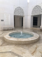 A serene courtyard with a circular water fountain at its center. The walls surrounding the courtyard are adorned with intricate Islamic geometric patterns in white. A stone bench is placed against one wall, next to an engraved plaque, adding to the tranquil atmosphere.