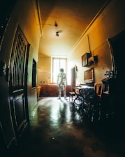 An astronaut stands in the middle of a dimly lit, vintage-looking room with a high ceiling. The walls and floors have an aged appearance, and the sunlight coming through the window highlights the figure in the space suit, creating an intriguing contrast between the old-fashioned interior and the futuristic attire.