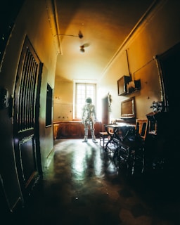 An astronaut stands in the middle of a dimly lit, vintage-looking room with a high ceiling. The walls and floors have an aged appearance, and the sunlight coming through the window highlights the figure in the space suit, creating an intriguing contrast between the old-fashioned interior and the futuristic attire.