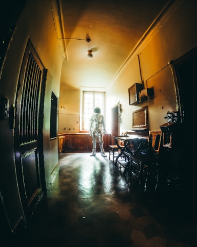 An astronaut stands in the middle of a dimly lit, vintage-looking room with a high ceiling. The walls and floors have an aged appearance, and the sunlight coming through the window highlights the figure in the space suit, creating an intriguing contrast between the old-fashioned interior and the futuristic attire.