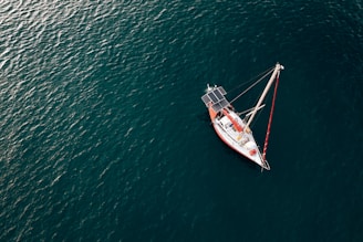 A lone sailboat with solar panels is floating on a vast expanse of deep blue ocean. The boat features a red and white color scheme and is calmly positioned, with visible masts and rigging.