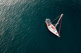 A lone sailboat with solar panels is floating on a vast expanse of deep blue ocean. The boat features a red and white color scheme and is calmly positioned, with visible masts and rigging.