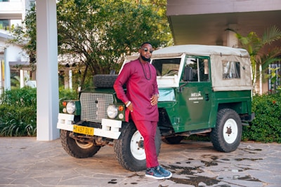 A man in a maroon traditional outfit stands confidently next to a vintage green off-road vehicle with a beige roof. The scene is set outdoors, surrounded by lush greenery and the corner of a building. The vehicle has a license plate and a sign reading 'Melia Serengeti Lodge'.