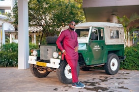 A man in a maroon traditional outfit stands confidently next to a vintage green off-road vehicle with a beige roof. The scene is set outdoors, surrounded by lush greenery and the corner of a building. The vehicle has a license plate and a sign reading 'Melia Serengeti Lodge'.