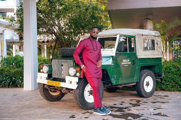 A man in a maroon traditional outfit stands confidently next to a vintage green off-road vehicle with a beige roof. The scene is set outdoors, surrounded by lush greenery and the corner of a building. The vehicle has a license plate and a sign reading 'Melia Serengeti Lodge'.