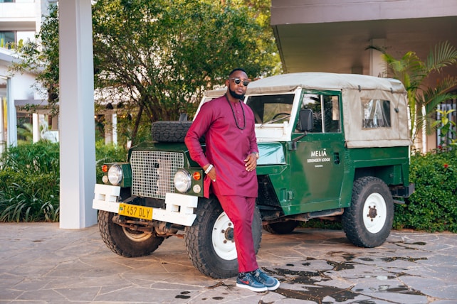A man in a maroon traditional outfit stands confidently next to a vintage green off-road vehicle with a beige roof. The scene is set outdoors, surrounded by lush greenery and the corner of a building. The vehicle has a license plate and a sign reading 'Melia Serengeti Lodge'.