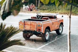 An orange vintage Land Rover parked on a paved area with a visible license plate. The surrounding environment includes some greenery and a road in the background. People are sitting in the distance near what looks like water containers.