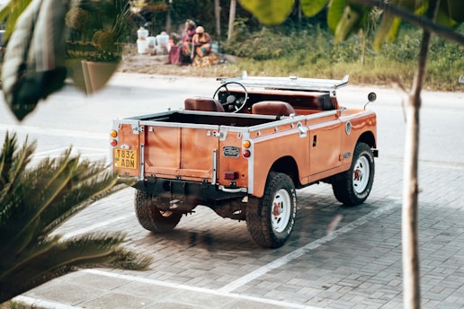 An orange vintage Land Rover parked on a paved area with a visible license plate. The surrounding environment includes some greenery and a road in the background. People are sitting in the distance near what looks like water containers.