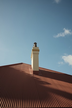 A chimney protrudes from the center of a brown corrugated metal roof under a clear blue sky. The shadows cast by the chimney and the texture of the roof add depth to the scene. A few white clouds are scattered in the sky.