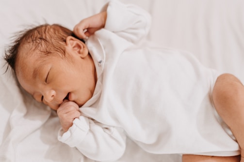 A newborn baby peacefully sleeps on a soft, white blanket while wearing a white onesie. The baby's hair is dark and slightly wet, and one hand is near the mouth, suggesting a soothing gesture.