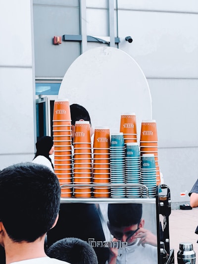 A stack of orange and teal disposable coffee cups is organized on top of a counter. The cups are branded with the word 'Bliss'. People are visible in the foreground and the reflection of one person can be seen on the stainless steel surface of the counter. The background consists of a white wall and a part of a door.