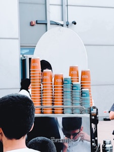 A stack of orange and teal disposable coffee cups is organized on top of a counter. The cups are branded with the word 'Bliss'. People are visible in the foreground and the reflection of one person can be seen on the stainless steel surface of the counter. The background consists of a white wall and a part of a door.