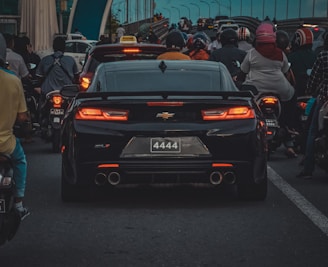 A black Chevrolet Camaro is stuck in traffic among a large number of motorcycles on a city road. The motorcycles and riders surround the car, filling the lanes behind and alongside it. The weather seems overcast, and the lighting suggests it might be evening or late afternoon with a generally bluish tint in the sky.