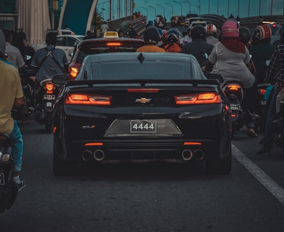 A black Chevrolet Camaro is stuck in traffic among a large number of motorcycles on a city road. The motorcycles and riders surround the car, filling the lanes behind and alongside it. The weather seems overcast, and the lighting suggests it might be evening or late afternoon with a generally bluish tint in the sky.
