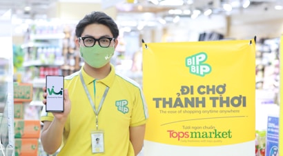 A person wearing a yellow uniform with green accents and a face mask stands in a store environment holding a smartphone displaying a logo. They have a lanyard with an ID card around their neck. To the side, a bright yellow banner promotes a shopping concept with the 'BIP BIP' logo and text in another language along with English, mentioning 'The ease of shopping anytime anywhere'. 'Tops market' is highlighted at the bottom of the banner.