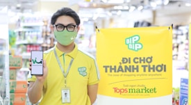 A person wearing a yellow uniform with green accents and a face mask stands in a store environment holding a smartphone displaying a logo. They have a lanyard with an ID card around their neck. To the side, a bright yellow banner promotes a shopping concept with the 'BIP BIP' logo and text in another language along with English, mentioning 'The ease of shopping anytime anywhere'. 'Tops market' is highlighted at the bottom of the banner.