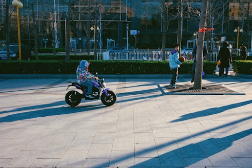 A child wearing a colorful jacket rides a small electric bike on a paved area in a park. Another child stands with a basketball next to a tree with a makeshift hoop. The scene takes place in an urban environment with buildings in the background and long shadows cast by the afternoon sun.