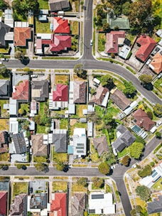 Aerial view of a residential neighborhood with neatly arranged houses, intersecting roads, and green yards. The houses have varied roof colors, including red, brown, and gray, surrounded by trees and lawns.