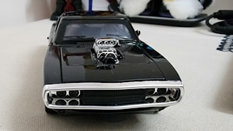 A black toy car with a shiny chrome supercharger on its hood is placed on a light-colored surface. The focus is on the front of the car, showcasing its grille and headlights. In the background, out-of-focus objects include a fluffy white pair of shoes or toys, a hint of a dark object, and some cables.