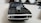 A black toy car with a shiny chrome supercharger on its hood is placed on a light-colored surface. The focus is on the front of the car, showcasing its grille and headlights. In the background, out-of-focus objects include a fluffy white pair of shoes or toys, a hint of a dark object, and some cables.