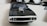 A black toy car with a shiny chrome supercharger on its hood is placed on a light-colored surface. The focus is on the front of the car, showcasing its grille and headlights. In the background, out-of-focus objects include a fluffy white pair of shoes or toys, a hint of a dark object, and some cables.