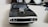 A black toy car with a shiny chrome supercharger on its hood is placed on a light-colored surface. The focus is on the front of the car, showcasing its grille and headlights. In the background, out-of-focus objects include a fluffy white pair of shoes or toys, a hint of a dark object, and some cables.