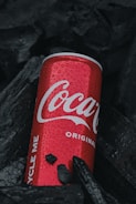 A red can with water droplets lies among dark charcoal pieces, its brand name prominently displayed in white lettering.