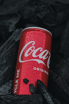 A red can with water droplets lies among dark charcoal pieces, its brand name prominently displayed in white lettering.