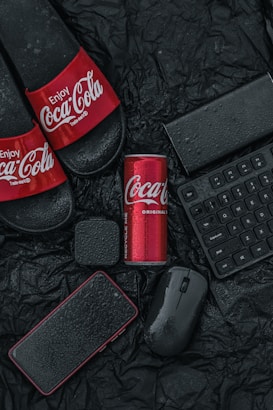 A collection of items including a pair of black slides with Coca-Cola branding, a red Coca-Cola can, a black mobile phone, a black wireless mouse, a wireless keyboard, and a small black speaker. These items are placed on a wrinkled black surface with water droplets visible on them, suggesting a wet or moist environment.