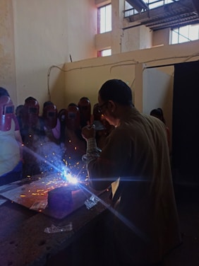 A group of people wearing protective face shields surrounds a person engaged in welding. Bright sparks and blue light emanate from the welding area, while smoke rises. The setting appears to be indoors with high ceilings and industrial elements, such as visible beams and windows.