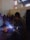 A group of people wearing protective face shields surrounds a person engaged in welding. Bright sparks and blue light emanate from the welding area, while smoke rises. The setting appears to be indoors with high ceilings and industrial elements, such as visible beams and windows.