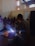A group of people wearing protective face shields surrounds a person engaged in welding. Bright sparks and blue light emanate from the welding area, while smoke rises. The setting appears to be indoors with high ceilings and industrial elements, such as visible beams and windows.
