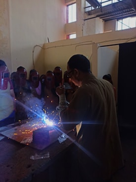A group of people wearing protective face shields surrounds a person engaged in welding. Bright sparks and blue light emanate from the welding area, while smoke rises. The setting appears to be indoors with high ceilings and industrial elements, such as visible beams and windows.