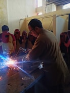A man is welding on a metal surface, producing bright sparks. He is surrounded by several people wearing red welding helmets. The setting appears to be an indoor workshop with walls showing signs of wear.