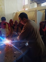 A man is welding on a metal surface, producing bright sparks. He is surrounded by several people wearing red welding helmets. The setting appears to be an indoor workshop with walls showing signs of wear.