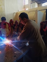 A man is welding on a metal surface, producing bright sparks. He is surrounded by several people wearing red welding helmets. The setting appears to be an indoor workshop with walls showing signs of wear.