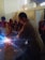 A man is welding on a metal surface, producing bright sparks. He is surrounded by several people wearing red welding helmets. The setting appears to be an indoor workshop with walls showing signs of wear.