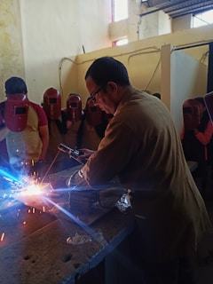 A man is welding on a metal surface, producing bright sparks. He is surrounded by several people wearing red welding helmets. The setting appears to be an indoor workshop with walls showing signs of wear.