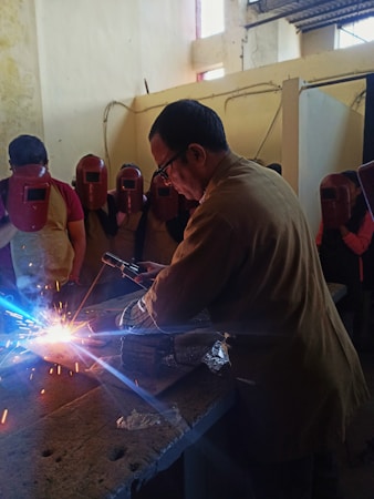 A man is welding on a metal surface, producing bright sparks. He is surrounded by several people wearing red welding helmets. The setting appears to be an indoor workshop with walls showing signs of wear.