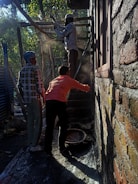 Three people are engaged in construction work, likely plastering or cementing a brick wall in an outdoor setting. Two men, one wearing a checkered shirt and a headscarf, and another in a bright orange shirt, are positioned near a third individual who is elevated on a scaffold. Light filters through surrounding trees, casting shadows and highlighting dust particles in the air.