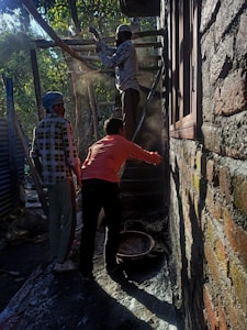 Three people are engaged in construction work, likely plastering or cementing a brick wall in an outdoor setting. Two men, one wearing a checkered shirt and a headscarf, and another in a bright orange shirt, are positioned near a third individual who is elevated on a scaffold. Light filters through surrounding trees, casting shadows and highlighting dust particles in the air.