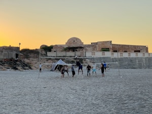 A group of people are playing volleyball on a sandy beach. The setting sun creates a warm glow in the sky, providing a calming backdrop. A rustic, ancient-looking building with stone walls is visible in the background. Some seating made from tires is arranged on one side.