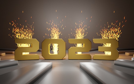 Large golden numbers depicting '2023' stand prominently against a background with bursts of fiery sparks emanating from behind them, creating a dynamic and celebratory atmosphere. The stage-like setting with geometric platforms adds depth to the scene.