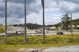 A construction site with heavy machinery including an excavator and trucks. The area is surrounded by a fence with a background of trees and a cloudy sky. Sparse vegetation and palm trees are visible in the foreground.