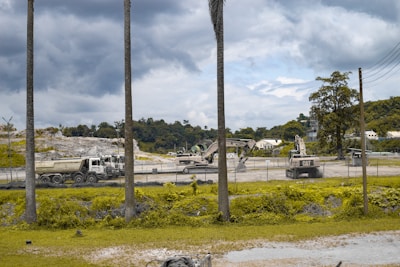 A construction site with heavy machinery including an excavator and trucks. The area is surrounded by a fence with a background of trees and a cloudy sky. Sparse vegetation and palm trees are visible in the foreground.