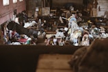 The image depicts a bustling indoor market or workshop filled with textiles and people. Various colorful fabrics and clothes are scattered throughout the room, with sewing machines visible in the background. People are engaged in activities related to the textile industry, possibly working or trading. The environment appears lively and somewhat chaotic, with a mixture of colors and textures.