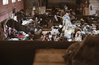 The image depicts a bustling indoor market or workshop filled with textiles and people. Various colorful fabrics and clothes are scattered throughout the room, with sewing machines visible in the background. People are engaged in activities related to the textile industry, possibly working or trading. The environment appears lively and somewhat chaotic, with a mixture of colors and textures.