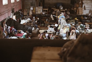 The image depicts a bustling indoor market or workshop filled with textiles and people. Various colorful fabrics and clothes are scattered throughout the room, with sewing machines visible in the background. People are engaged in activities related to the textile industry, possibly working or trading. The environment appears lively and somewhat chaotic, with a mixture of colors and textures.